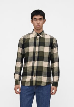 Jeune homme portant une chemise à carreaux verte, noire et beige à manches longues avec un jean bleu, debout devant un fond blanc uni.