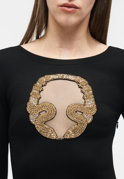 Γυναίκα που φοράει μαύρο τοπ με διάφανο ύφασμα στο στήθος με σχέδιο χρυσού φιδιού από χάντρες.