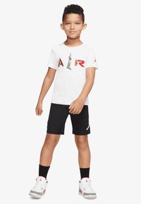 Weißes T-Shirt mit rotem "AIR"-Text und Grafik, schwarze Shorts, weiße Sneaker mit schwarzen Akzenten und einem dekorativen Muster auf den Schuhen.