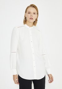 Witte blouse met lange mouwen en hoge kraag, voorzien van een knoopsluiting aan de voorkant, subtiele schouderuitsnijdingen en manchetten. Gladde stof met een losse pasvorm.