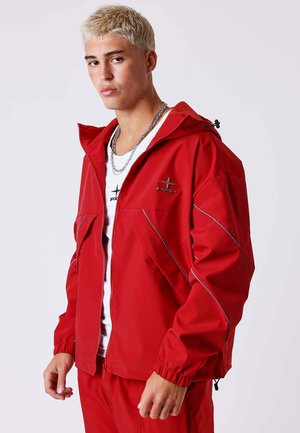 Jeune homme aux cheveux blonds courts portant une veste zippée rouge et un pantalon assorti par-dessus un t-shirt blanc, debout devant un fond uni.