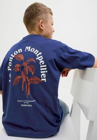 Tume sinine lühikeste varrukatega t-särk, millel on pruun botaaniline trükikujundus ja tekst "Le Ponton Montpellier" ning "Studio Dept." seljapool.