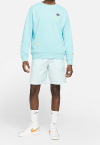 Felpa di cotone azzurro chiaro con logo Nike, abbinata a pantaloni corti verde menta con ricami sottili e sneaker bianche con accenti arancioni.