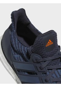 Gros plan d'une basket Adidas bleu marine avec une texture tricotée, une semelle en mousse, des lacets et un logo Adidas orange sur la languette.