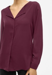 Blusa bordeaux a maniche lunghe con scollatura a V e un sottile pattina anteriore. Tessuto liscio con una leggera lucentezza e polsini svasati.