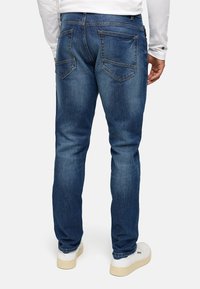 Mørkeblå denimjeans, rett passform, med to baklommer og mild fading. Brukt med en hvit langermet skjorte og hvite sko.