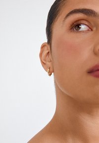 Pendientes de aro dorados con un diseño retorcido, que cuentan con una superficie pulida y un cierre seguro. El modelo tiene una complexión media.