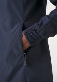 Mörkblå jacka med elastiska muddar, en zipficka på sidan och slät, vattenavvisande tyg med en elegant design.