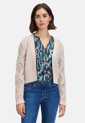 Beige gestrickte Strickjacke mit Lochmuster, verkürztem Design und langen Ärmeln. Getragen über einem blauen gemusterten Oberteil und kombiniert mit Jeans.