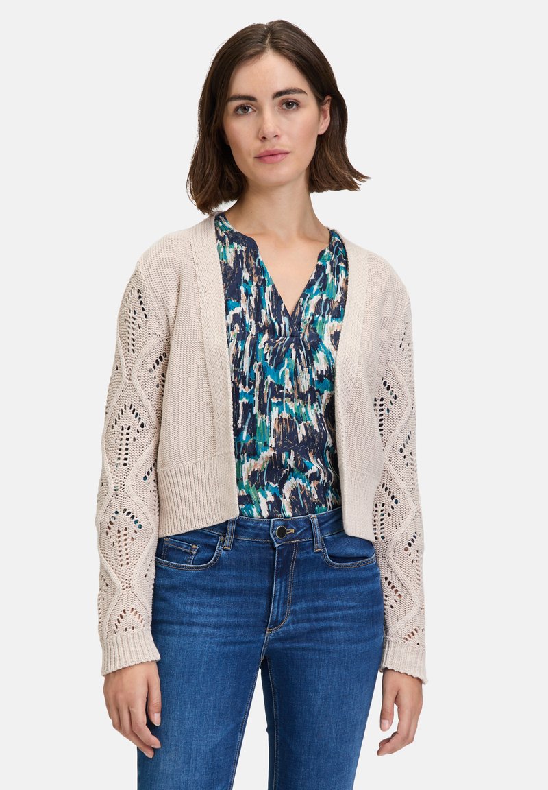 Beige gebreide cardigan met opengewerkte patroond, cropped ontwerp en lange mouwen. Gelaat over een blauwe gebloemde top en gecombineerd met denim jeans.