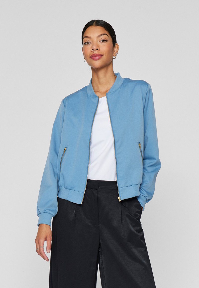 Svetlo modra bomber jakna z rebrastim ovratnikom, zadrgo, stranskimi žepi in elastičnimi manšetami. Oblečena preko bele srajce in črnih hlač.