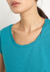 Tyrkysové tričko s krátkými rukávy a strukturovaným výstřihem do lodi. Tkanina se zdá být měkká a lehká, s jemným melírovaným vzorem.