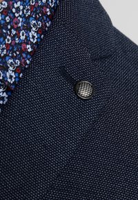 Mörkblå texturerad blazer med slag, en knapp med rutmönster och en glimt av en skjortkrage med blommönster.