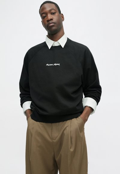 MustMustere mustahil täiesti vabalt lõigatud üherealine mustatsärk, millel on valge "Maison" tikand, raglanvarrukad ja ümar kaelus, mille all on valge särk.