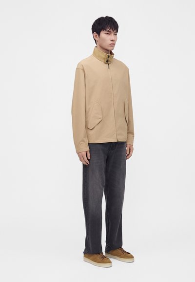 Homme debout portant une veste beige à col montant, un jean foncé et ample, et des chaussures décontractées marron sur un fond blanc uni.