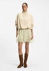 Mujer con blusa beige de cuello alto, minifalda de encaje color crema y botas de ante marrón hasta media caña, de pie frente a un fondo blanco.