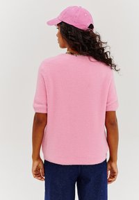 Chlpatý ružový sveter s krátkymi rukávmi a okrúhlym výstrihom, doplnený o tmavomodré nohavice. Modelka má na sebe zladenú ružovú čiapku.