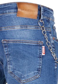 Kék farmernadrág bézs bőr címkével, ezüst láncos részletekkel és kontrasztos narancssárga varrással. Hátsó zsebében logós címke található.