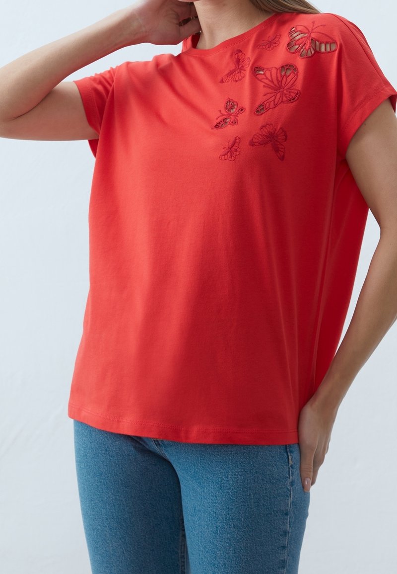 Femme portant un haut rouge à manches courtes avec des découpes en forme de papillon sur l'épaule, associé à un jean bleu, main sur le cou et la hanche.