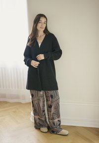 Femeie stand în interior, îmbrăcată cu un cardigan lung negru, pantaloni largi cu model și pantofi bej, pe podea din lemn deschis.