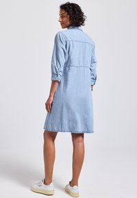 Robe en denim bleu clair avec une coupe décontractée, devant à boutons et manches retroussées. Elle présente un ourlet mi-long et des détails de couture subtils.
