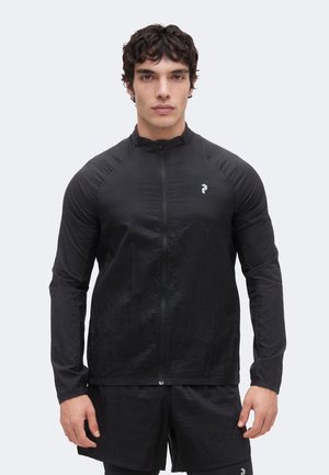 Młody mężczyzna z krótkimi ciemnymi włosami, ubrany w czarną sportową kurtkę na zamek i pasujące szorty, stojący na jednolitym tle.