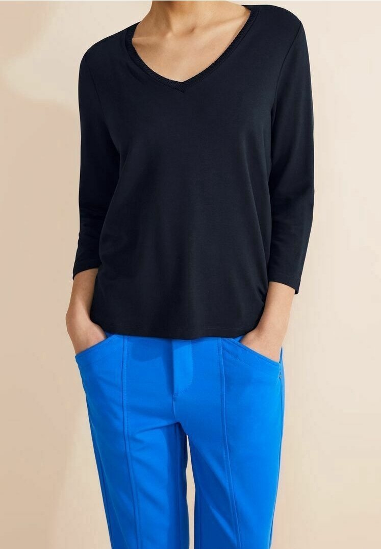 Personne portant un haut noir à manches longues avec un col en V et un pantalon bleu, les mains dans les poches, debout devant un fond beige.