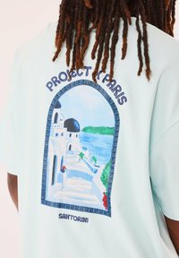 Camiseta de color azul claro con un diseño impreso de un paisaje de Santorini que incluye arquitectura blanca, cúpulas negras y acentos verdes. El texto sobre el diseño dice "PROYECTO PARÍS."