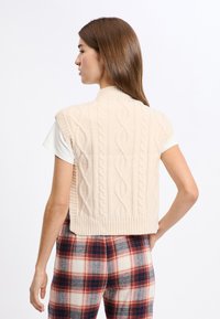 Ärmelloser Strickpullover in sanftem Beige mit hohem Kragen, der ein Kabelmuster und einen gerippten Saum aufweist. Getragen über einem weißen T-Shirt und karierten Hosen.