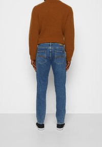 Mörkblå denimjeans med rak passform, fem fickor och synlig sömnad bak. Till dessa bärs en brun ribbad tröja och svarta skor.