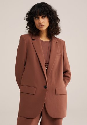 Frau mit lockigem, dunklem Haar, die einen braunen, übergroßen Blazer und passende Hosen vor einem einfarbigen beigefarbenen Hintergrund trägt.