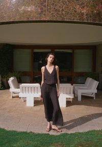 Young woman in black sleeveless jumpsuit and sandals stands on patio with white outdoor furniture and glass doors behind her.