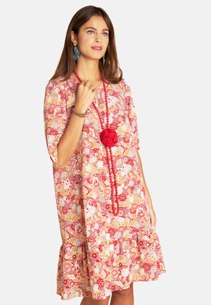Rotes Blumenkleid mit kurzen Ärmeln, gerafftem Saum und kunstvollen Mustern. Akzentuiert mit einer roten Blume und einer Perlenkette.