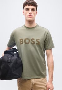 Νεαρός άνδρας που φοράει πράσινο ελιάς μπλουζάκι "BOSS" και μπεζ παντελόνια, κρατώντας μια μαύρη τσάντα duffel στο αριστερό του χέρι.