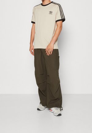 T-shirt beige avec bordure noire, arborant le logo Adidas sur le côté gauche de la poitrine. Pantalon cargo vert olive avec multiples poches. Chaussures de sport grises.