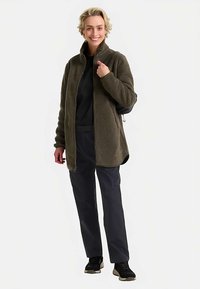 Veste en polaire vert olive avec col montant, fermeture éclair et poches latérales, associée à un top noir et un pantalon foncé, portée avec des baskets.