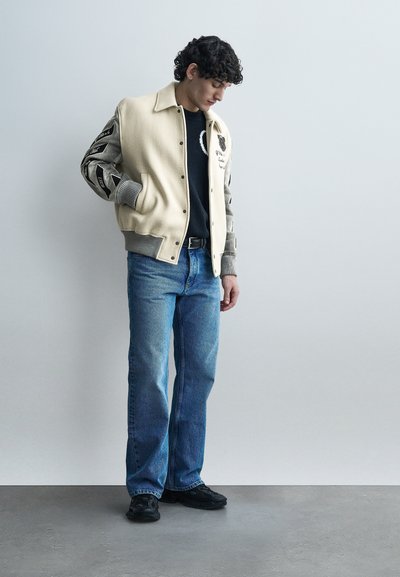 Νεαρός άνδρας που φοράει μπεζ και γκρι varsity μπουφάν, μαύρο πουλόβερ, μπλε τζιν και μαύρα αθλητικά παπούτσια, στέκεται με τα χέρια στις τσέπες μπροστά σε λευκό τοίχο.