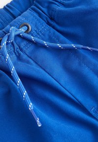 Blå badshorts med elastisk midja och snörning. Snörningen är flätad, med plastspetsar. Slät tygstruktur.