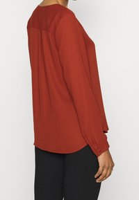 Blouse couleur rouille à coupe décontractée, manches longues et poignets boutonnés. Comprend un ourlet arrondi et un tissu doux et léger.