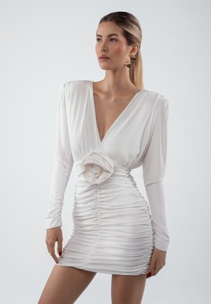 Kobieta z eleganckim kucykiem, ubrana w białą, marszczoną mini sukienkę z długimi rękawami, głębokim dekoltem w kształcie litery V oraz dużym kwiatowym zdobieniem w talii.