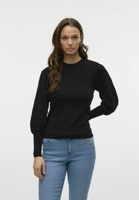 Zwarte pullover trui met gestructureerde pofmouwen, ronde halslijn en aangesloten manchetten. Gecombineerd met lichtblauwe denim jeans.