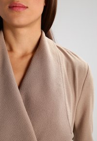 Nais yang mengenakan blazer berbahan tekstur beige dengan kerah lebar, memperlihatkan leher, bagian bawah wajah, dan bahu di latar belakang yang polos.
