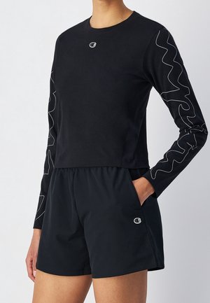 Personne portant un haut noir à manches longues avec des motifs de lignes blanches ondulées sur les manches et un short noir assorti avec un logo sur la poche gauche de la jambe.