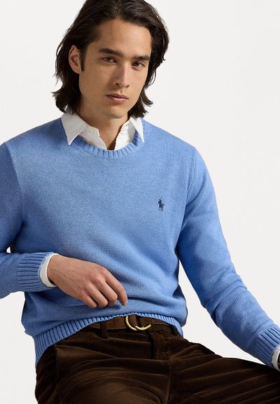 Pull en tricot bleu clair avec un col rond et des poignets côtelés, arborant un petit logo sombre sur la poitrine. Modèle portant un pantalon en velours côtelé marron.