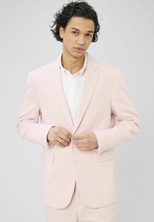 Homme aux cheveux bouclés et foncés boutonnant une veste de costume rose clair sur une chemise blanche, sur un fond uni.