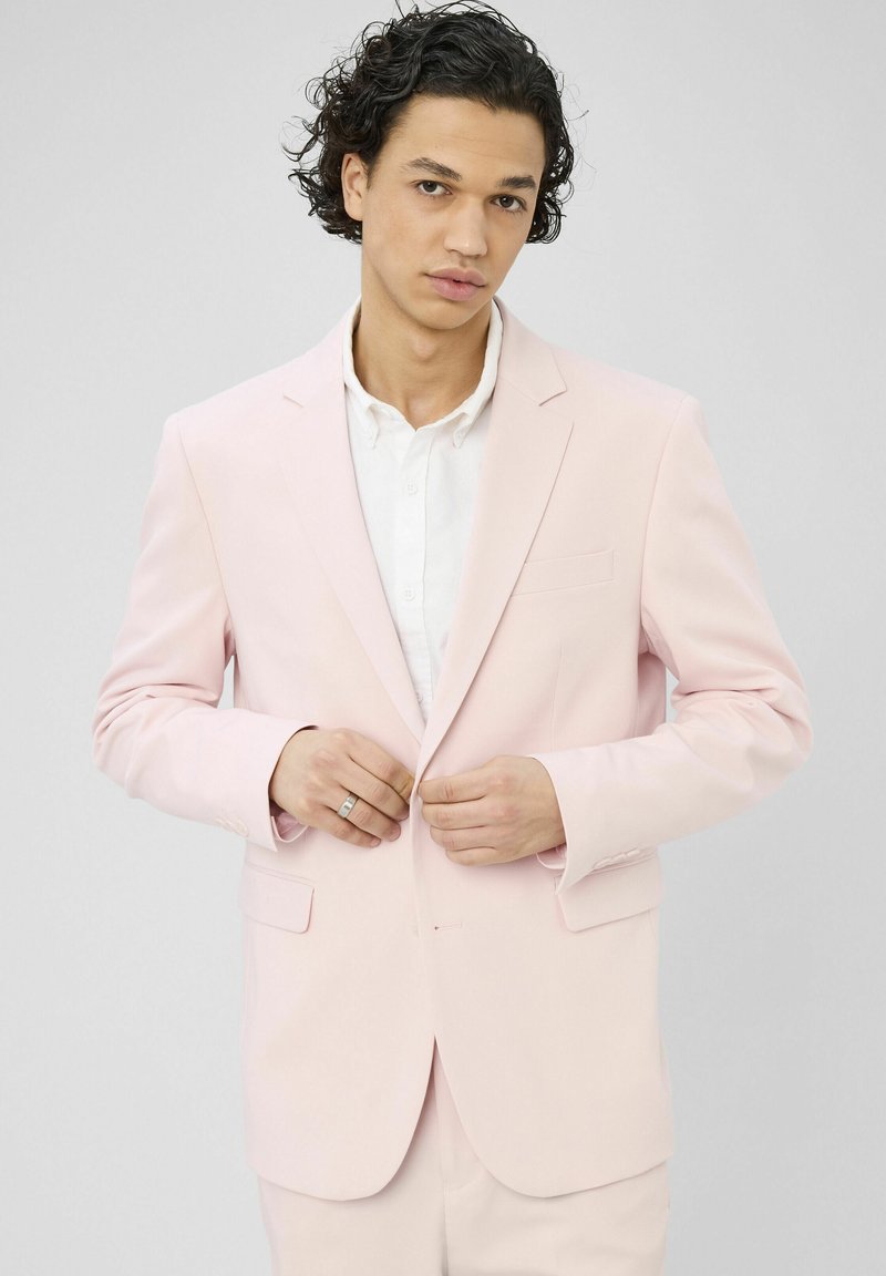 Homme aux cheveux bouclés et foncés boutonnant une veste de costume rose clair sur une chemise blanche, sur un fond uni.