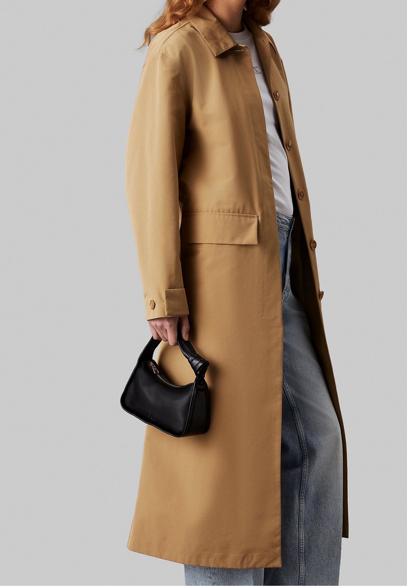Femme portant un long manteau beige par-dessus une chemise blanche et un jean, tenant un petit sac à main noir devant un fond gris uni.