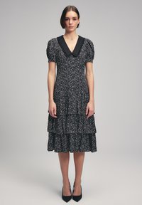 Zwarte jurk met bloemenprint, tiered ontwerp, puffmouwen en een zwarte kraag. Geweven stof met kleine witte stippen.