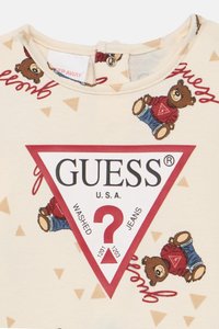 Μπεζ ύφασμα με τρίγωνο λογότυπο Guess με κόκκινες περιγραφές, σχέδιο με αρκουδάκια που φορούν κόκκινα πουλόβερ και μπλε τζιν, καθώς και μικρά μπεζ τρίγωνα.