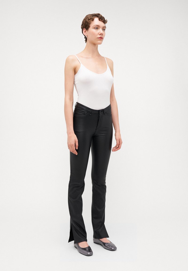 Schwarze, taillierte Hosen mit seitlichen Schlitzen, kombiniert mit einem weißen ärmellosen Body. Das Model trägt metallische Ballerinas und steht vor einem schlichten Hintergrund.